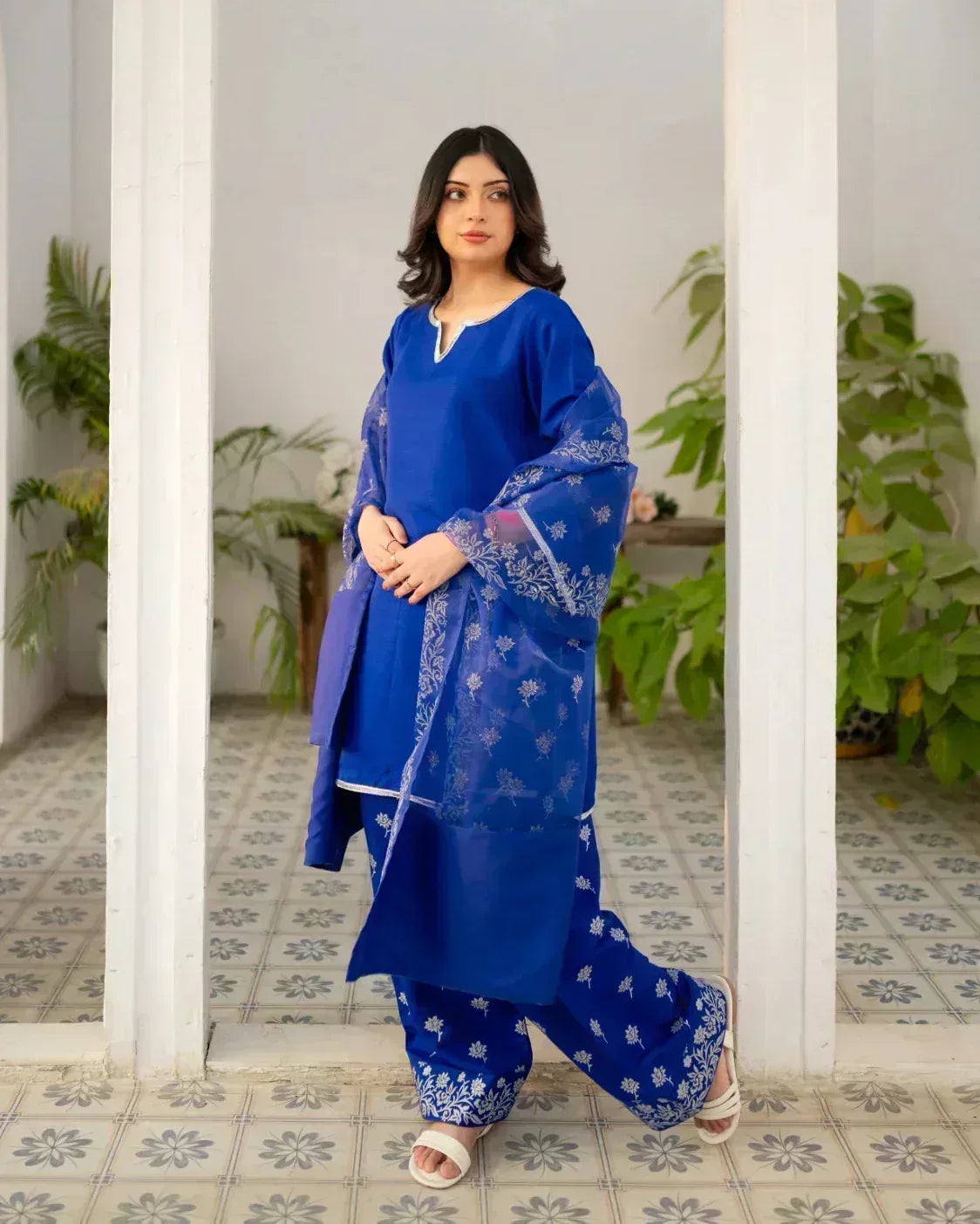 Woman in a royal blue traditional farshi shalwar suit standing in a decorated room with plants.