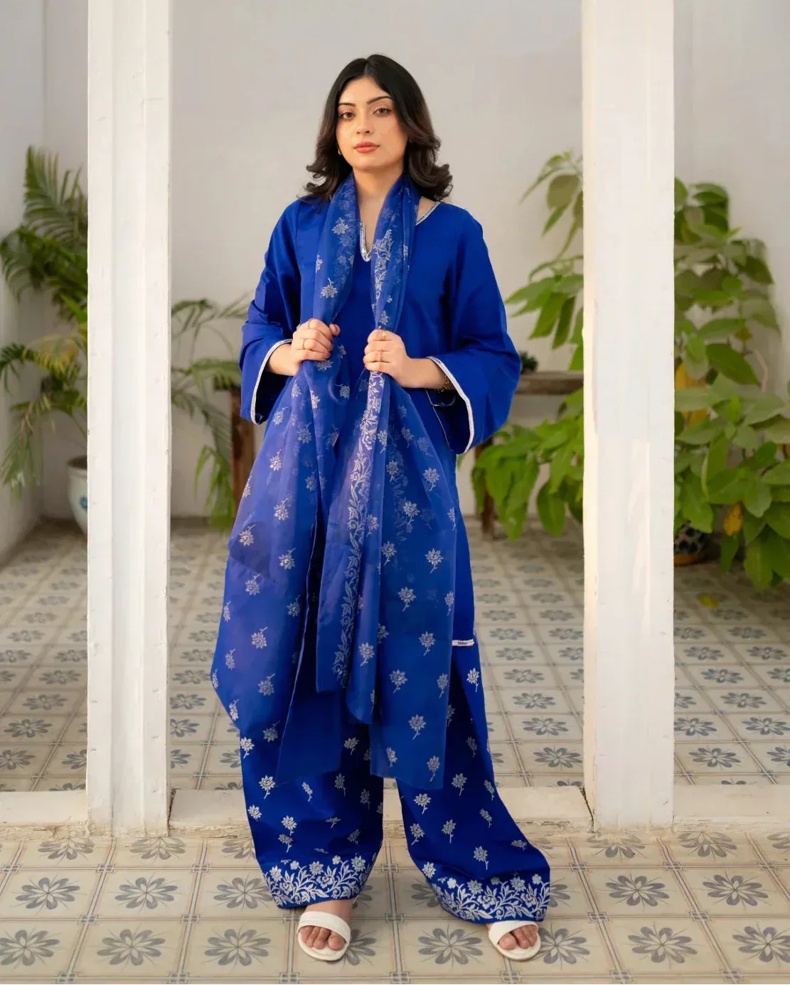 Woman wearing a royal blue traditional farshi shalwar suit with white patterns in an indoor setting with plants.