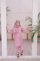 Child in a pink floral outfit standing in a room with plants and a tiled floor.