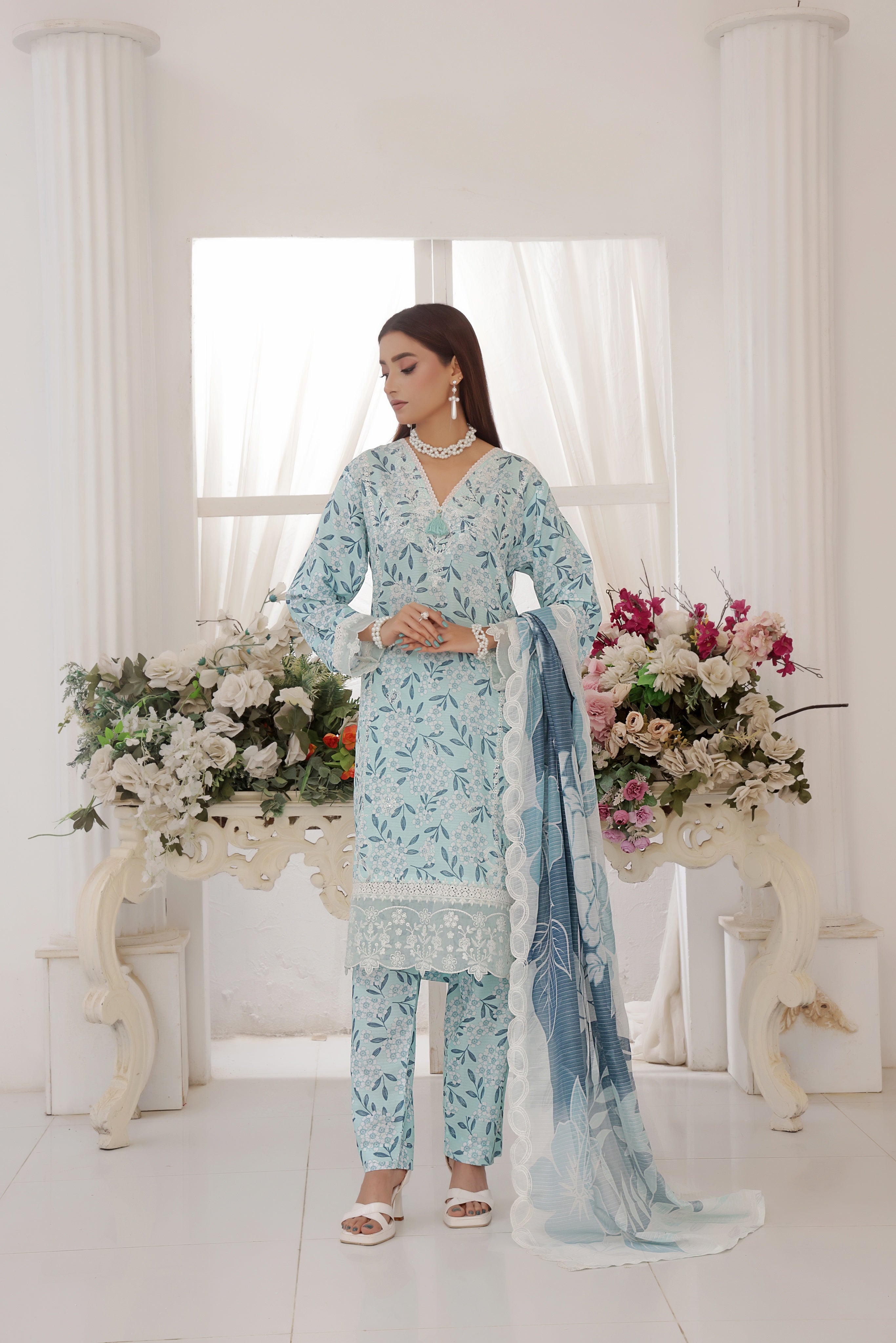 ZARNAZ - Woman in a light blue floral traditional outfit standing in a decorated room with flowers.