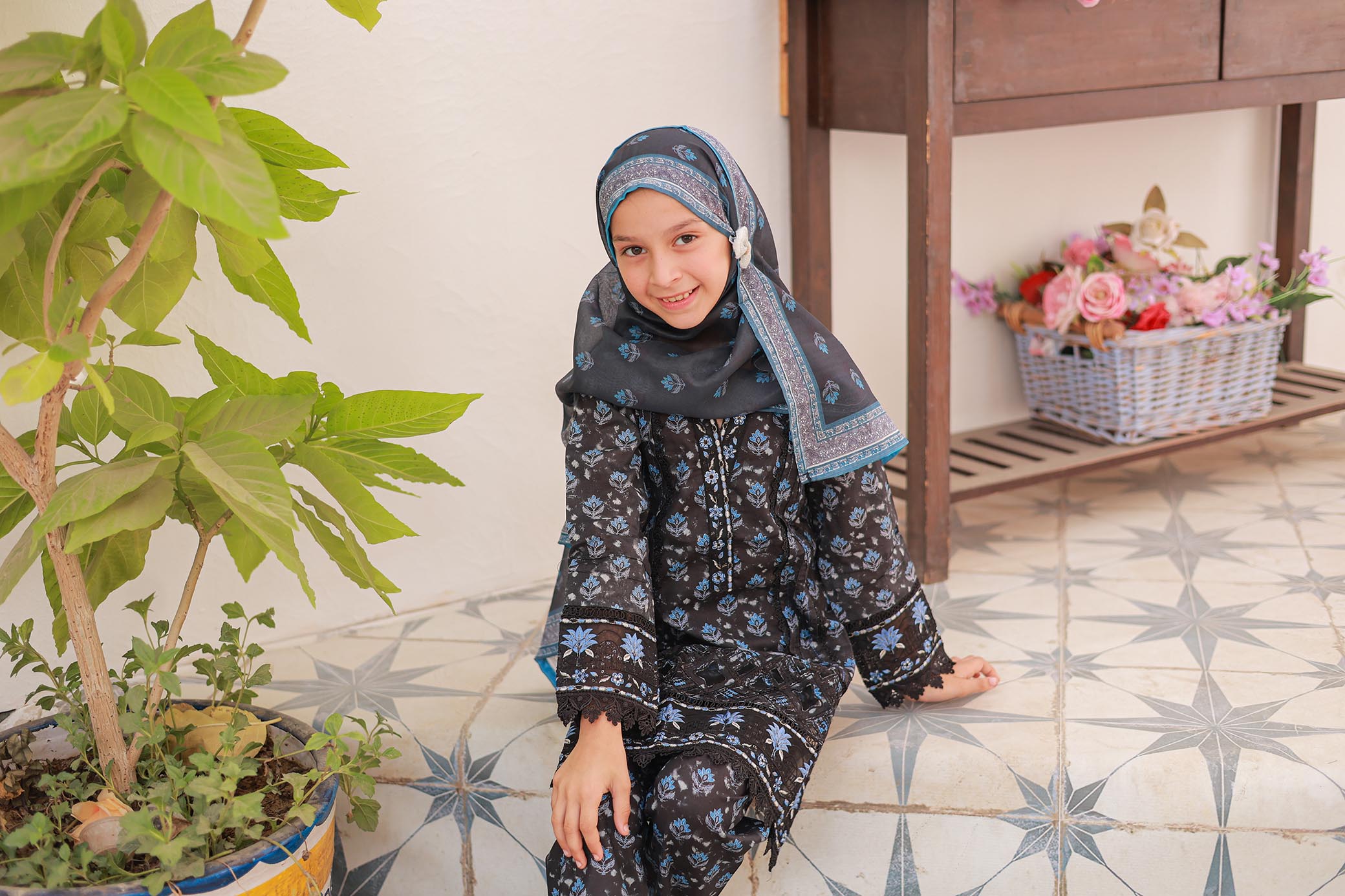 Child wearing a black floral outfit with a headscarf, sitting on a tiled floor next to a plant.