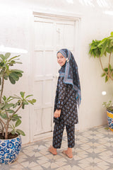 Woman wearing a floral outfit and headscarf standing in a room with plants and a white door.