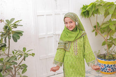 Child wearing a green traditional outfit with a headscarf, standing indoors with plants in the background.