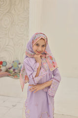 Woman wearing a light purple dress with floral patterns and a matching headscarf in a room with a floral-patterned wall.