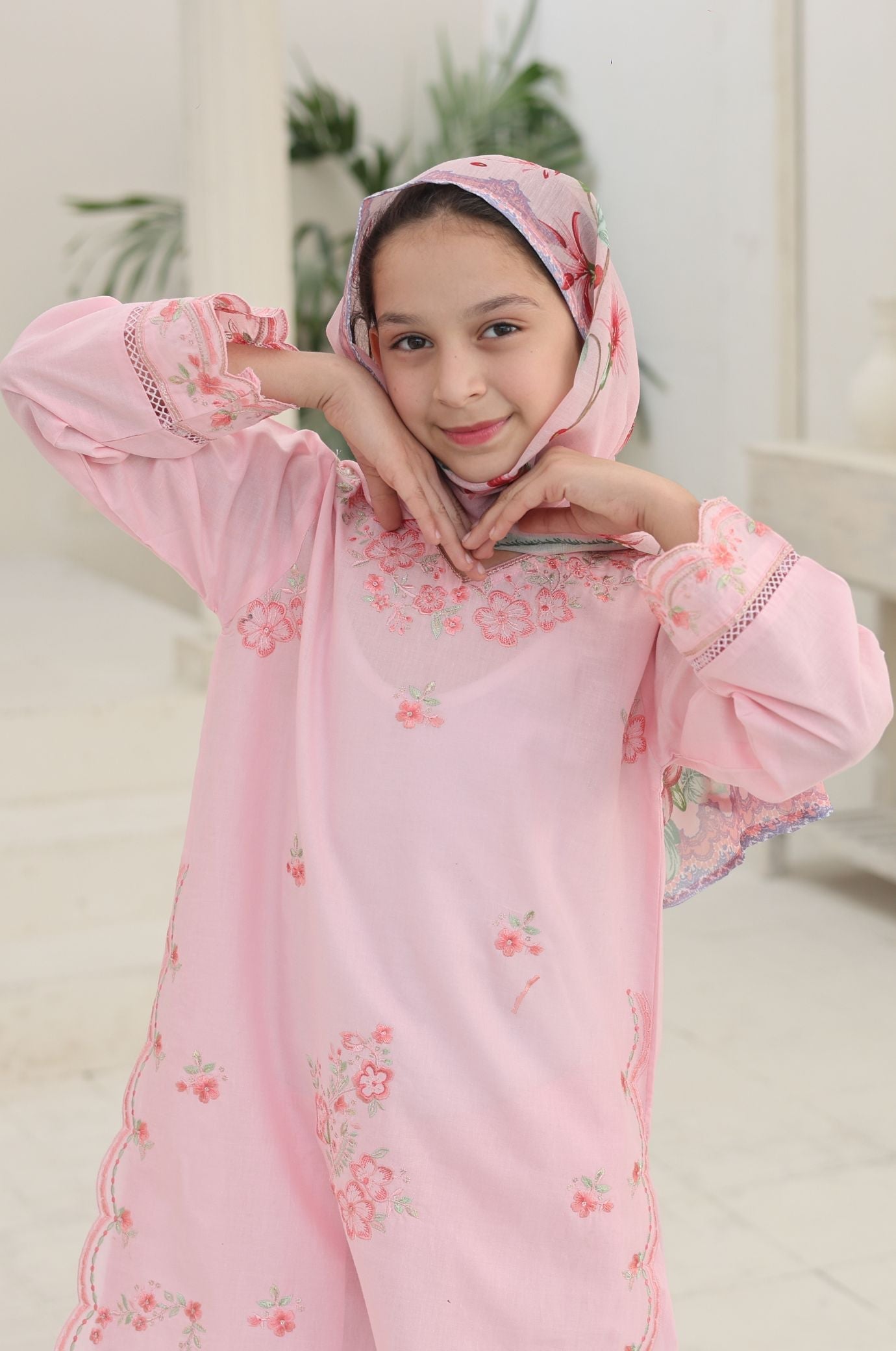 Young girl wearing a pink floral dress and headscarf indoors.