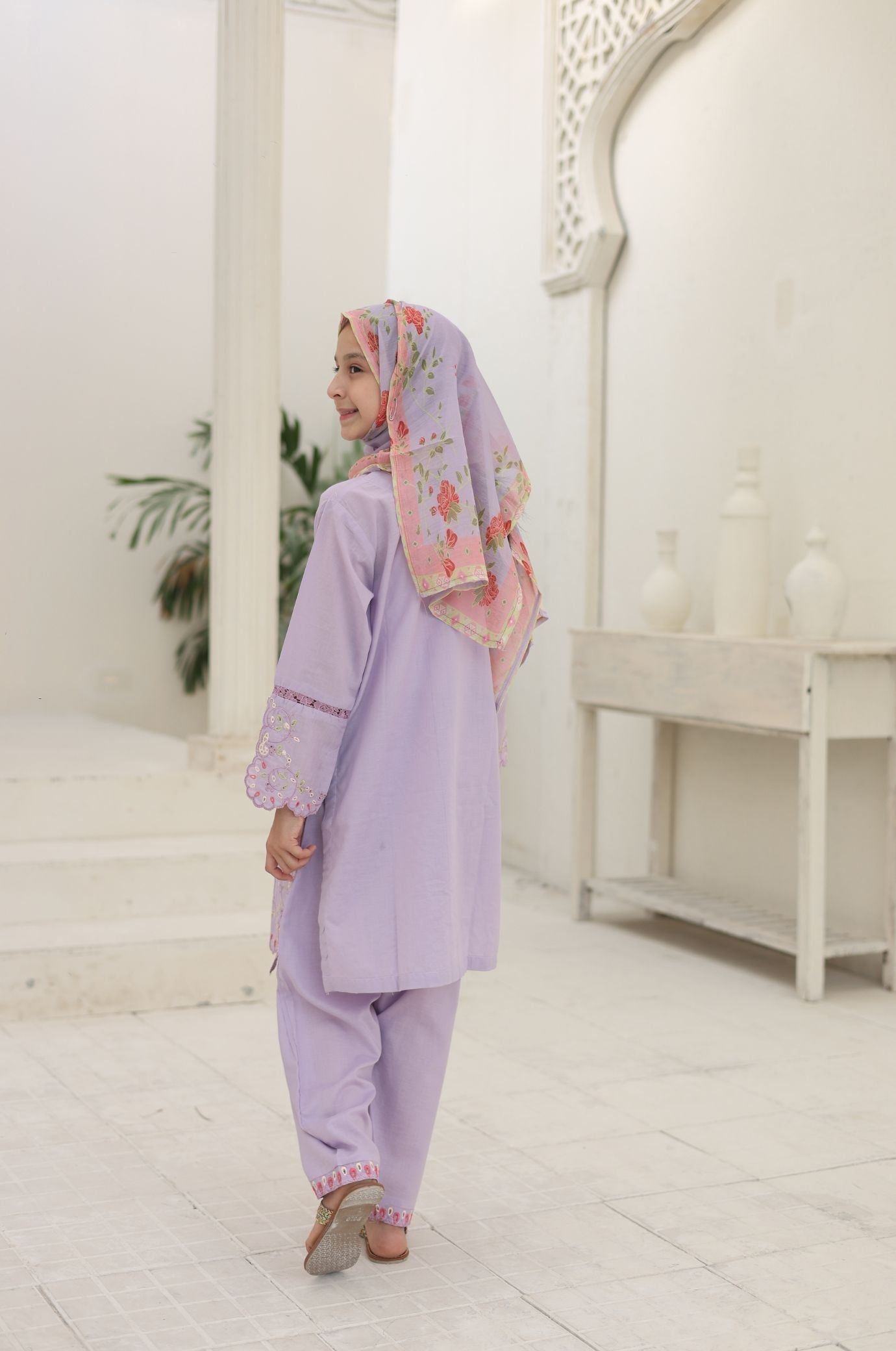 Woman in a lavender outfit with a floral headscarf standing in a white interior setting.