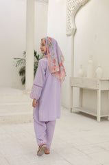 Woman in a lavender outfit with a floral headscarf standing in a white interior setting.