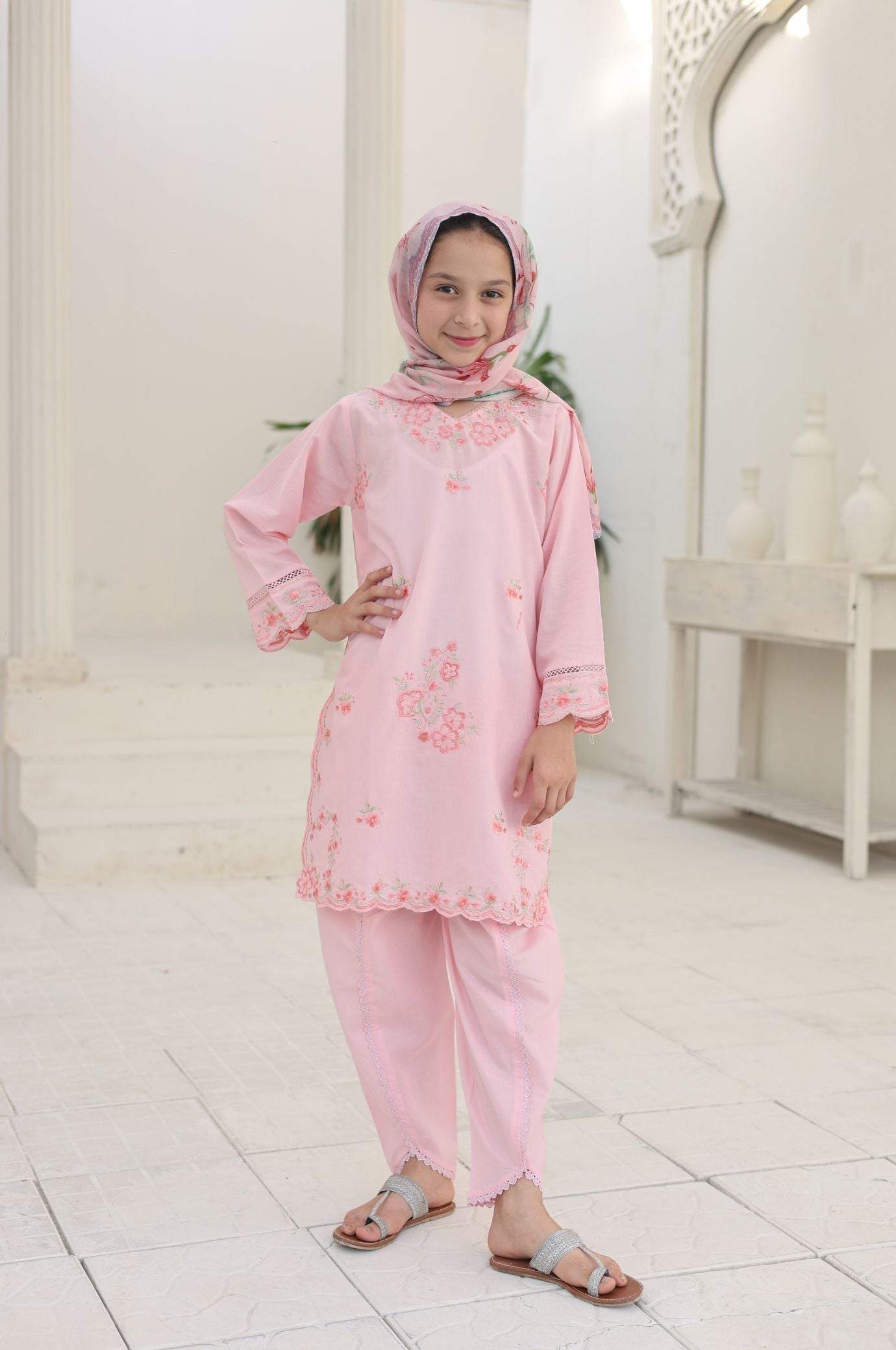 Child wearing a pink traditional outfit with a headscarf in a white room.