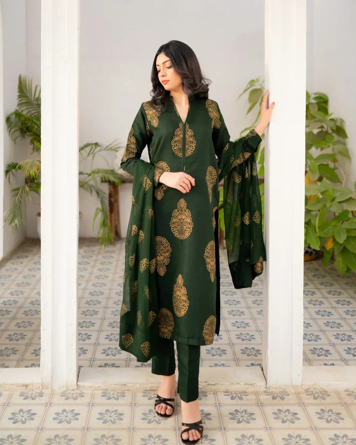 Woman wearing a bottle green traditional three piece suit with gold patterns in a room with plants and white pillars.