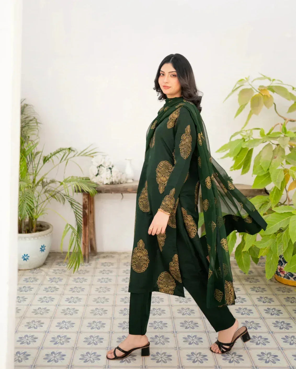 Woman wearing a bottle green traditional outfit with gold patterns in a room with plants and a table.