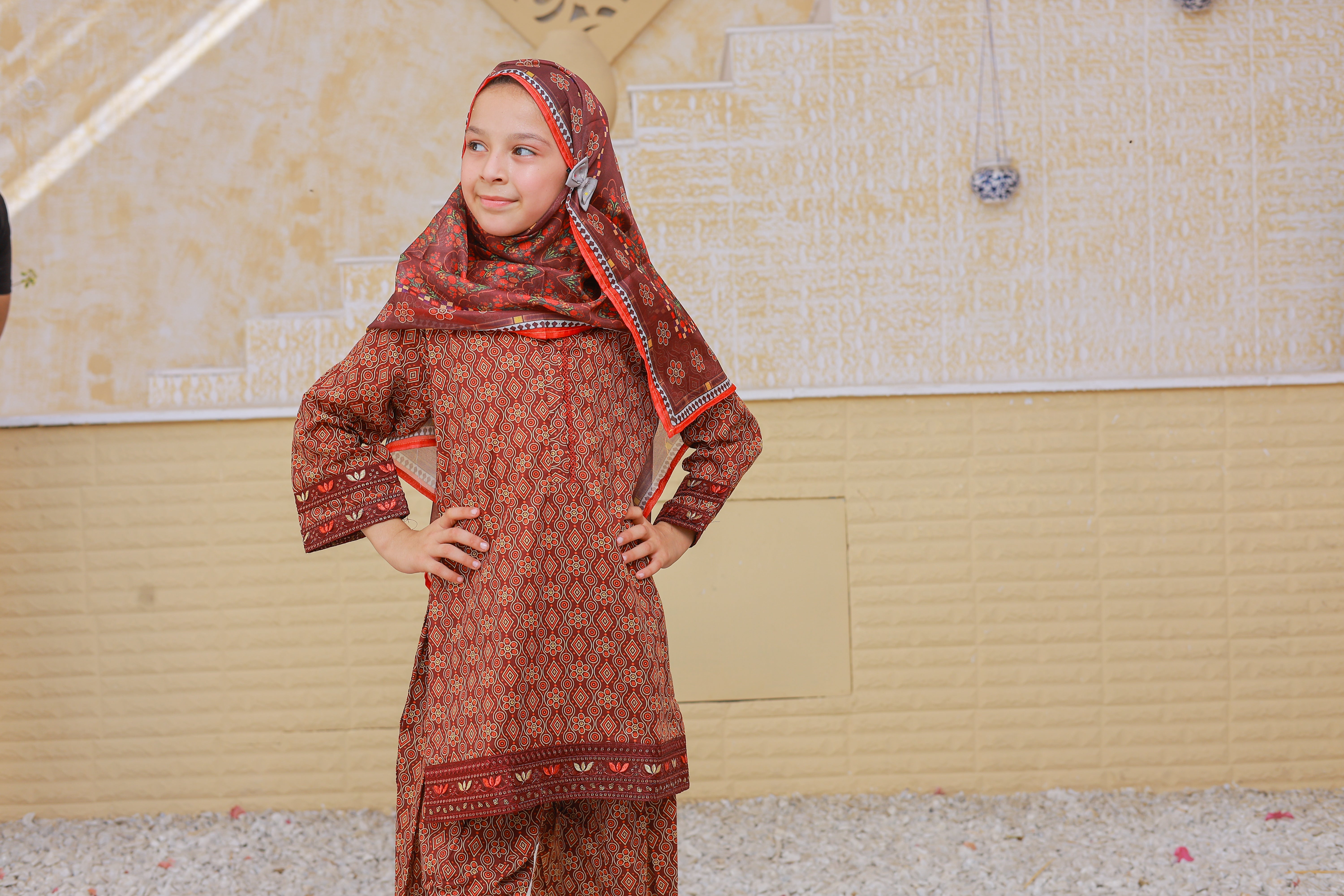 Young girl wearing a traditional red and patterned outfit with a headscarf in an indoor setting.