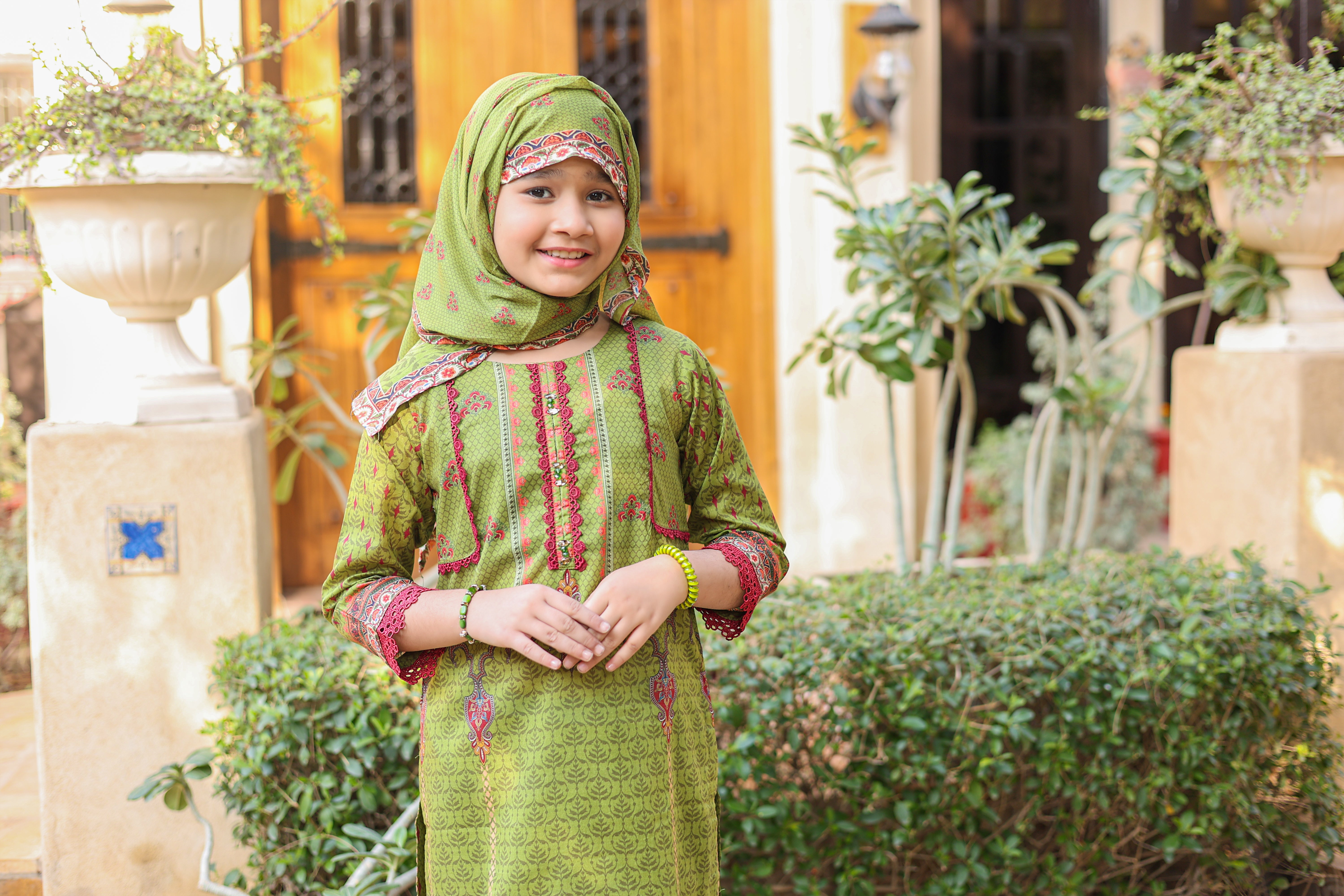 Young girl in a green traditional outfit standing outdoors with plants and a building in the background