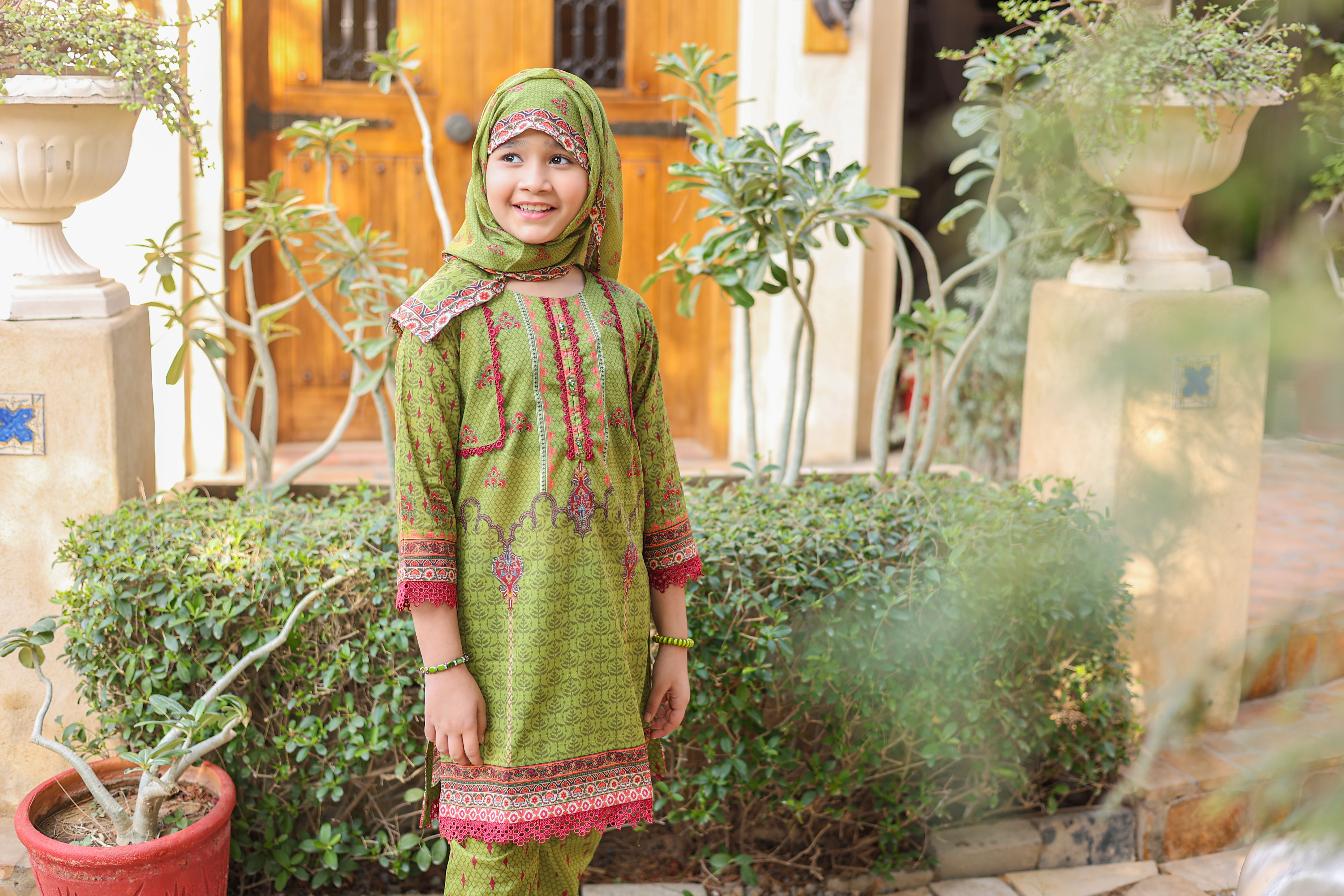 Woman wearing a pink floral dress and headscarf indoors with a decorative wall and plant in the background.