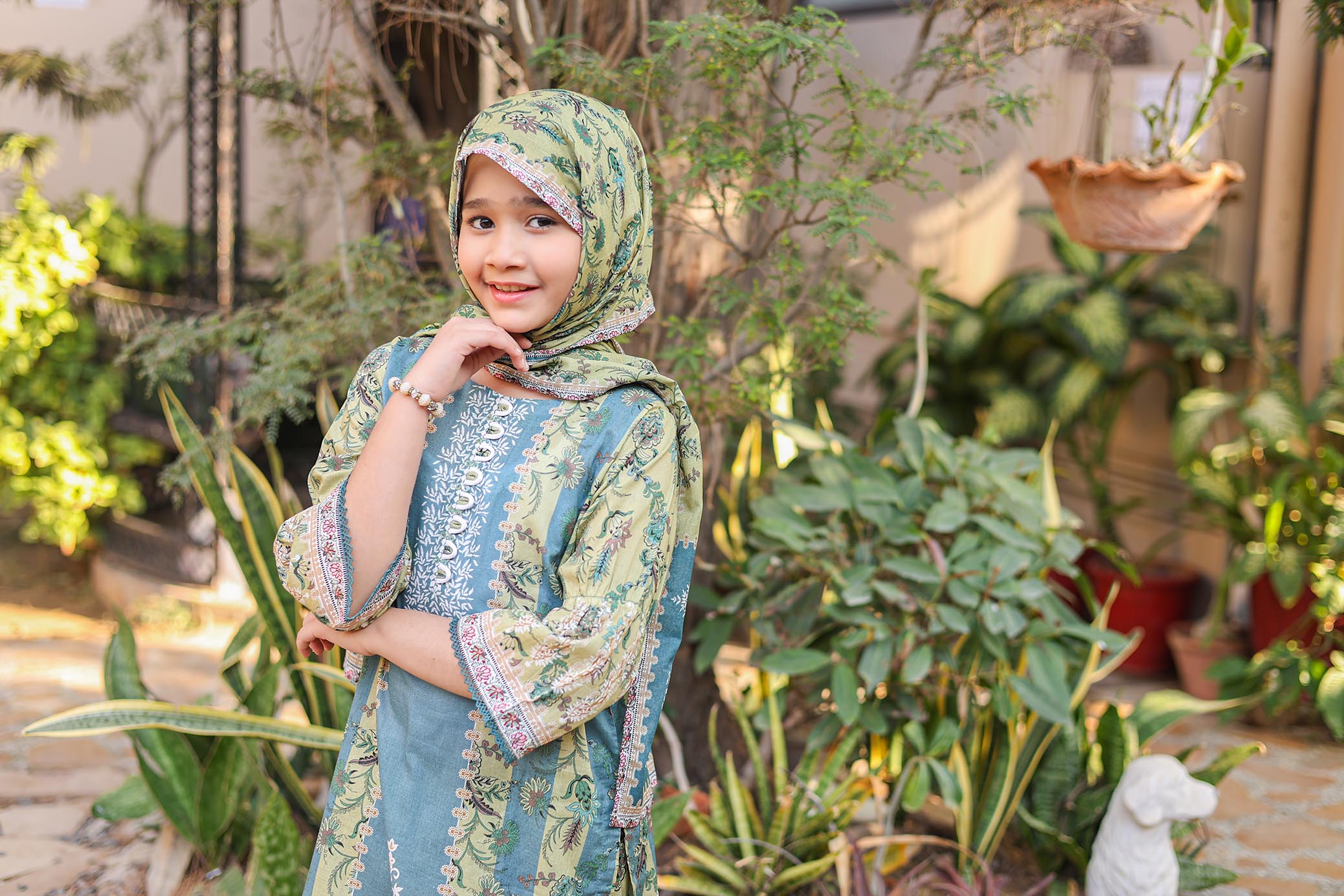 Young girl in a patterned dress and hijab standing in a garden with plants around.