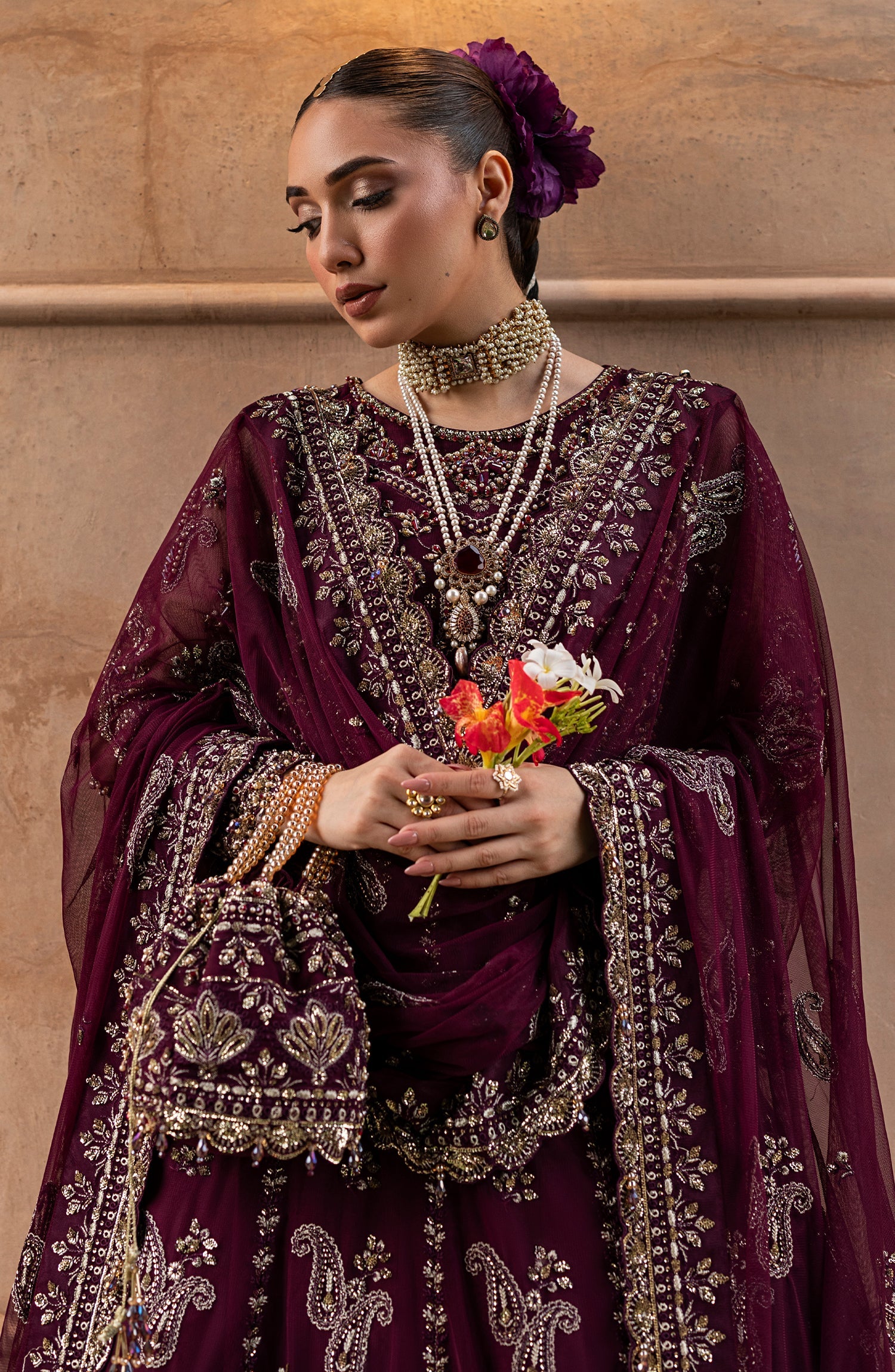 Woman in a traditional embroidered outfit holding flowers against a beige wall.