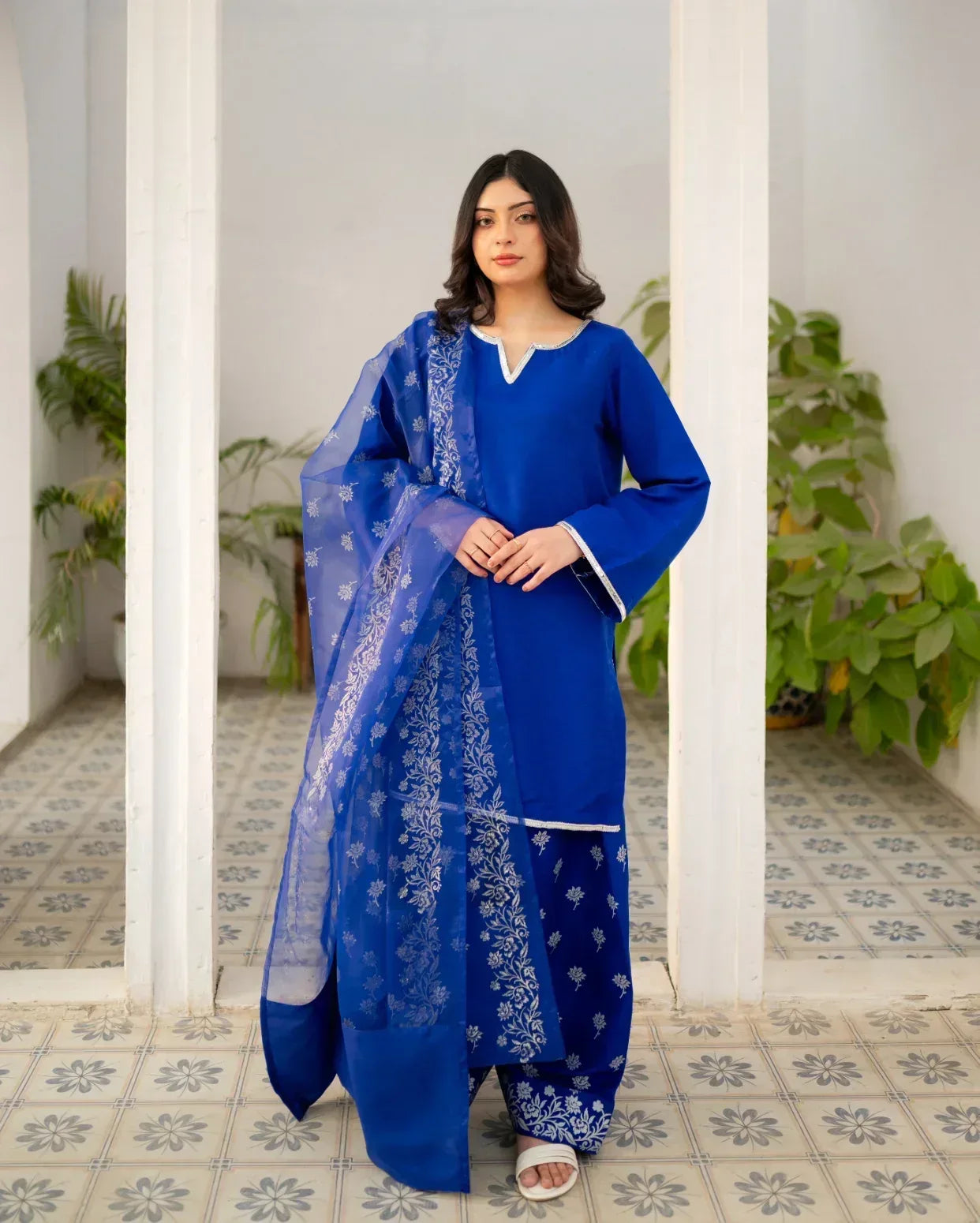 Woman wearing a royal blue traditional farshi shalwar suit with a white border in an indoor setting with plants.
