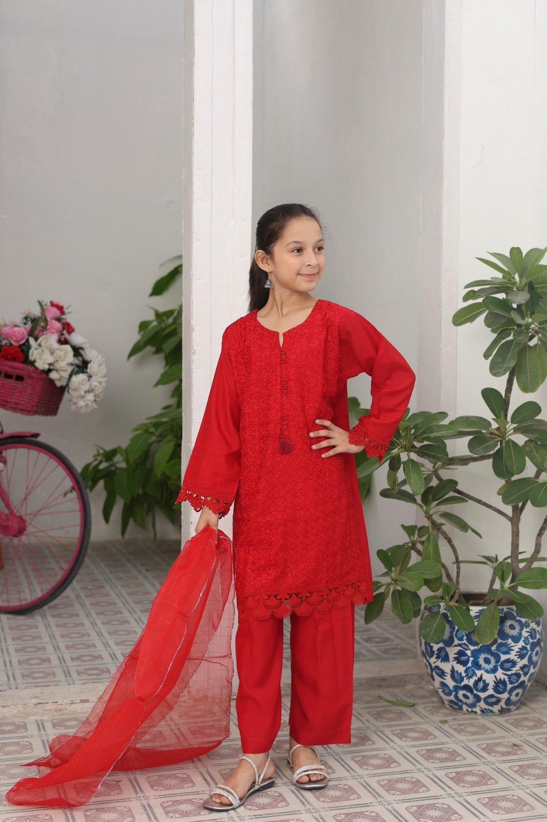 Young girl in a red traditional outfit standing indoors with plants and a bicycle in the background.