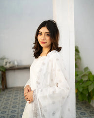Woman in a white kurta set standing against a light-colored wall with plants in the background.